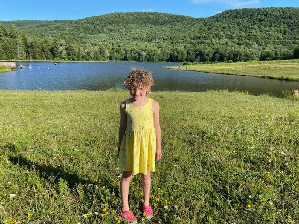 A young girl wearing a yellow dress stands in a green field with Colgate Lake in the background, which is one of the best family-friendly beaches in the Catskills.