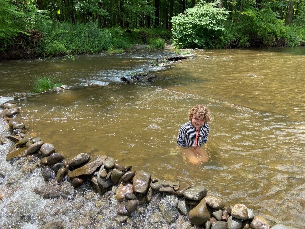 A young child wearing a striped swim suit kneels in a large river off the Comeau Property Trail, one of the most kid-friendly hikes in the Hudson Valley.