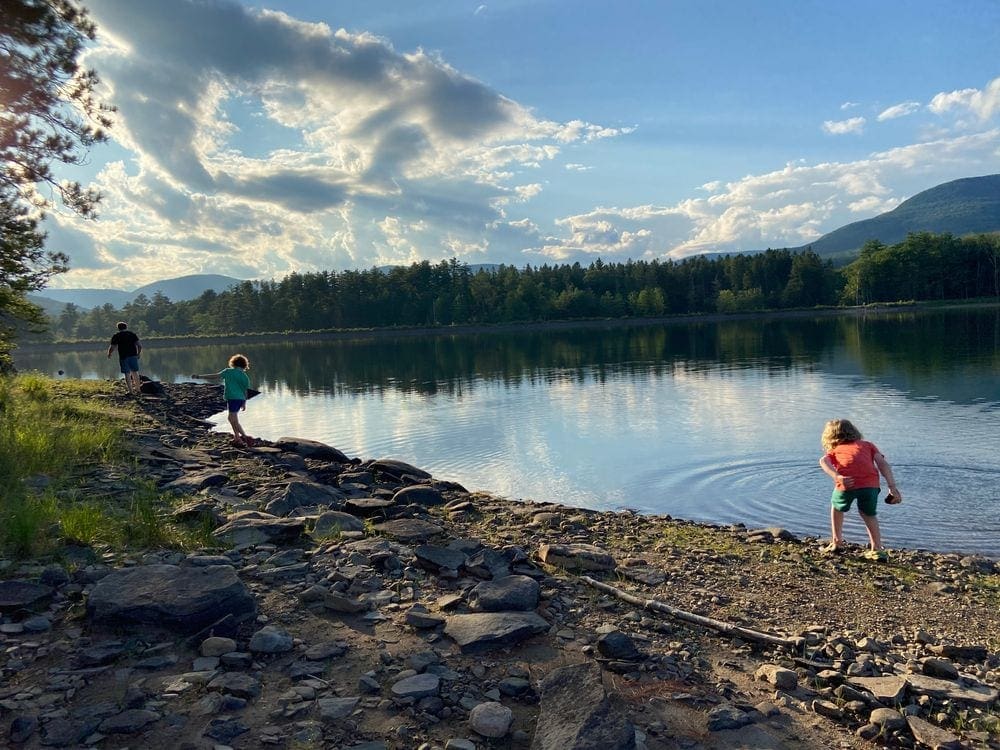 A man and two children are shown wandering the shower of Cooper Lake Reservoir with evergreen trees dotting the otherside of the bank.