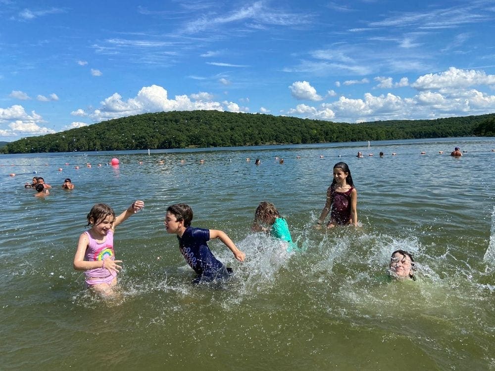 Five kids splash wildly in the water at East Beach. Other swimmers dot the background, as a line of lush green trees flank the other bank of the lake.