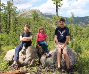 Three young kids sit along two large boulders with several green trees behind them.