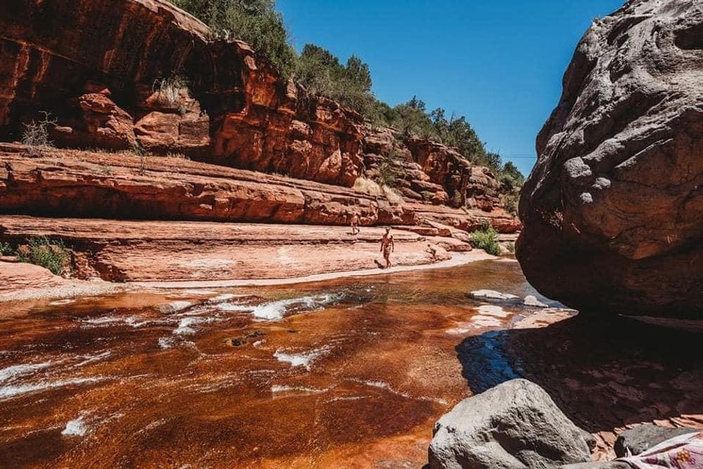 A sweeping view of a stream cutting through the red rocks at Slide Rock State Park, while a solo walker enjoys the sun,
