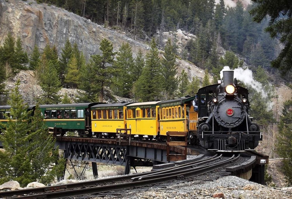 A train with three yellow cars and two green cars zooms into focus while going around a bend. Taking the Georgetown Loop Railroad is one of the best drives to see fall colors in Colorado with kids.