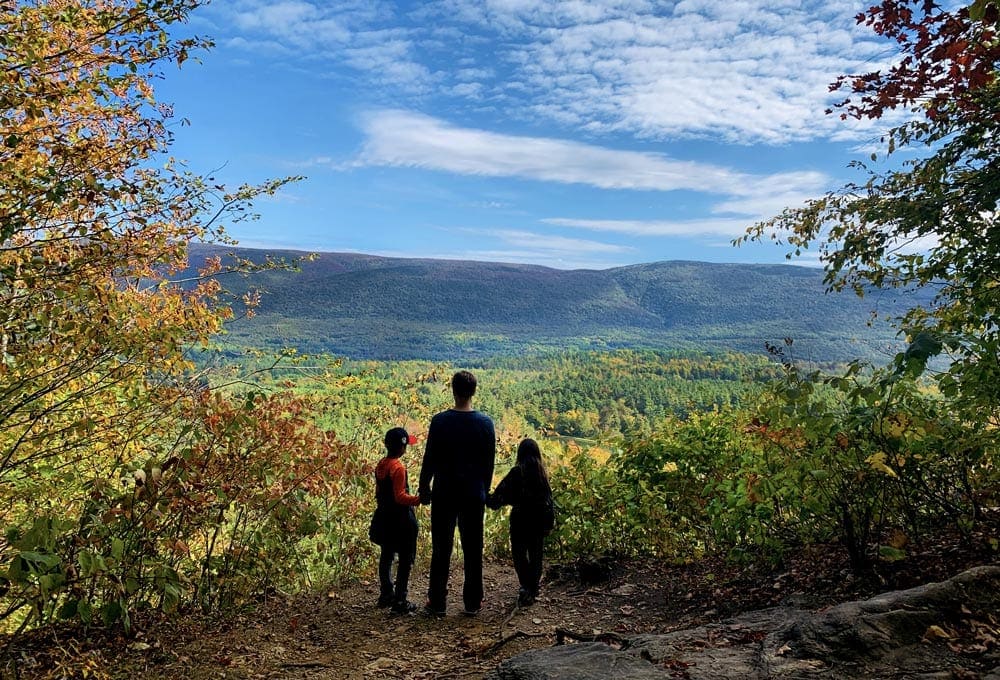 An adult holds the hands of two small children while standing on the edge of a cliff overlooking an incredible fall scene on a brilliantly sunny day in Litchfield, one of the Best Cute Towns To Visit With Kids Near NYC.