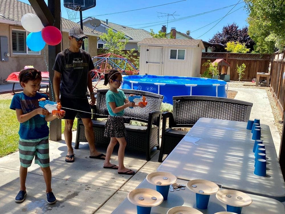 A dad looks on as his young son and daughter compete in a game where the squirt guns they are holding need to spray water to knock down blue solo cups.