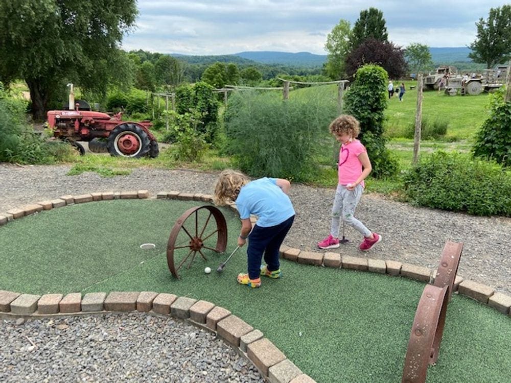 A young child attempts to get a mini-golf ball through a wheel as part of the putting green, while an older sibling looks on. If in the Catskills with kids, try mini-golfing at Kelder Farms.