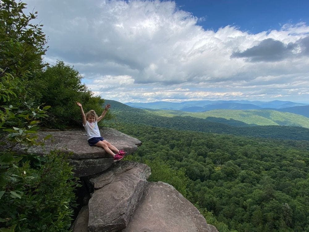 A young girl sits on a high rock ledge with her arms excitedly in the air. Around her, lush green trees dot the expansive view of the Catskill Mountains.