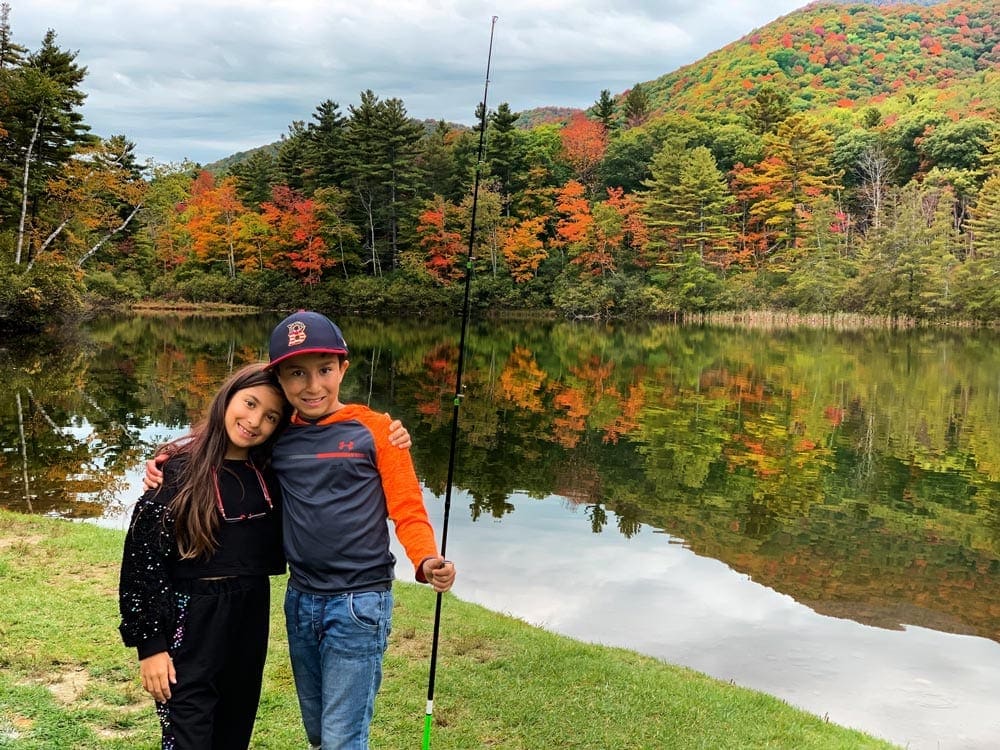 Sister and brother stand close together, while the boy holds a fishing pole. Behind them is a glimmering lake flanked on the far bank with a full array of striking fall colors.