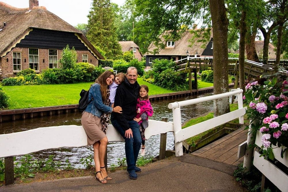 A family of four stands against a white fence along a Dutch canal with a traditional Dutch-styled home behind the canal.