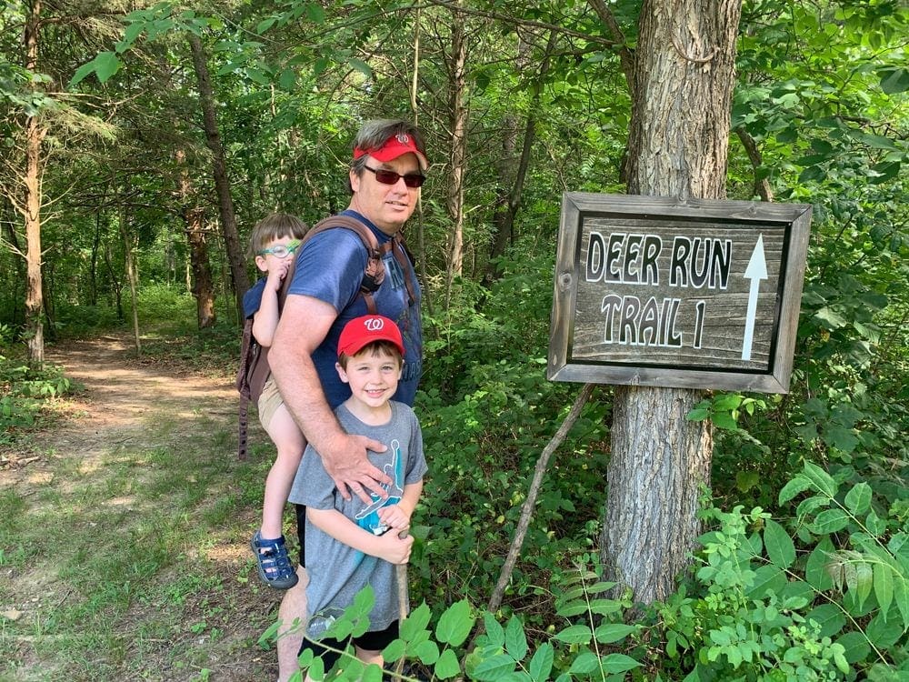 A dad stands need a trail sign that reads "Deer Run Trail 1", while his older son stands in front of him and the younger son sits in a back carrier on his back in one of the best places to visit in the Shenandoah River Valley with Kids.