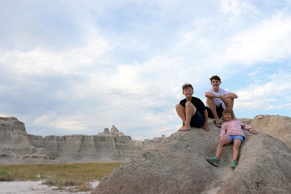 Three kids sit smiling atop a large rock in Badlands National Park, one of the best stops on a socially distant vacation with kids.