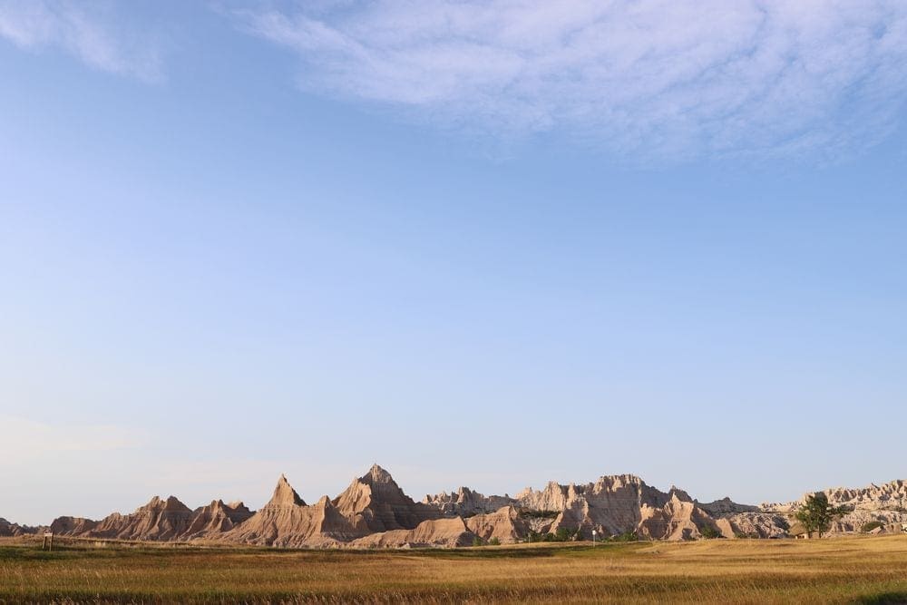 An expansive view of the Badlands National Park, in the foreground is a grassy field, with large rock formations in the background.