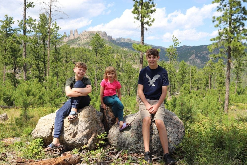 Three young kids sit along two large boulders with several green trees behind them.