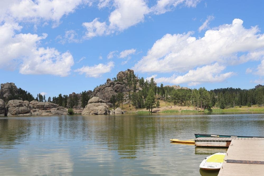 A pituresque lake is surrounded by large rock formations, green trees, and a dock. Spending time on the water is a great way to embrace a socially distant vacation with kids.
