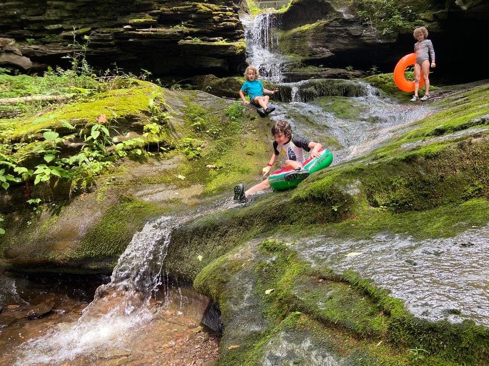 A young boy on a plastic inner tube rushes down a small waterfall as his two siblings watch.