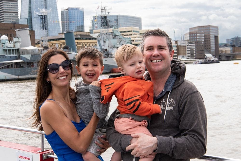 A family of four smiles at the camera, while large boats float down the Thames River in the background.