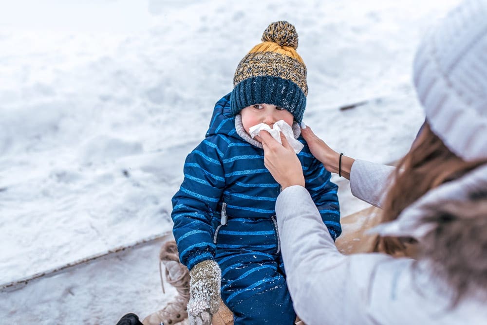 A mother helps her young child blow their nose outside. The mother wears a white coat and a white winter hat, while her child wears a blue striped snow suit and yellow and blue winter hat.