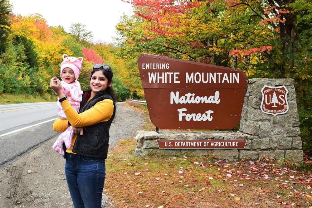 A mom holds her infant daughter in front of the White Mountain National Forest sign on a brilliant fall afternoon, one of the best places for fall foliage in New England.