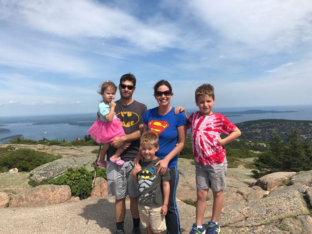 A family of five stands on top of the rocks with an expansive view of Acadia National Park behind them.