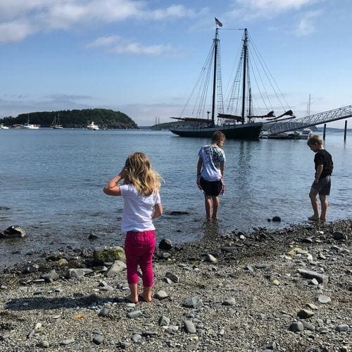 Three kids stand playing in the water with a large ship in the background.