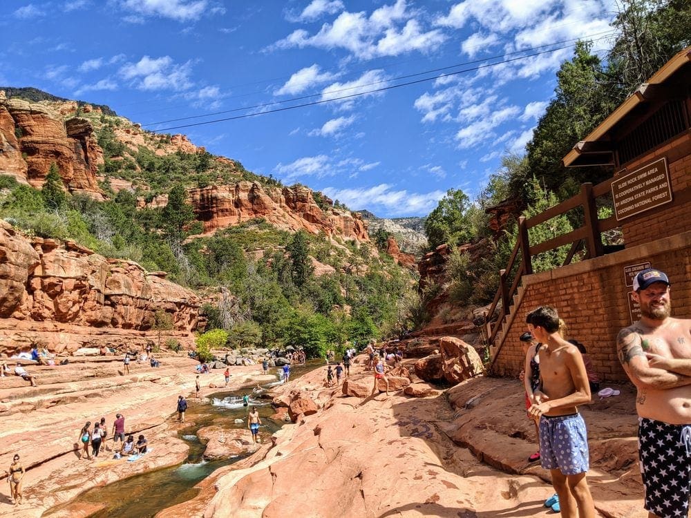 An expansive view of Slide Rock State Park, featuring several hikers and swimmers around a flowing stream through iconic red desert rocks.