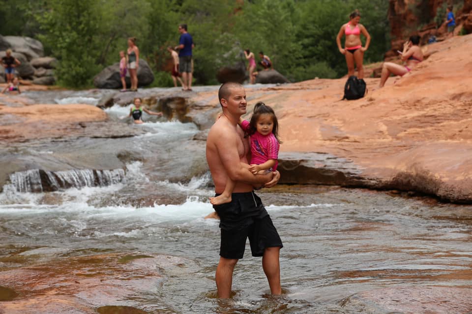 A dad carries his toddler daughter across a streem in Slide Rock State Park. Other swimmers dot the background.