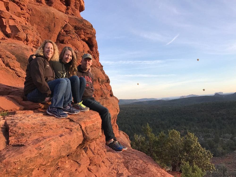 A family of three sits on one of Sedona's red rocks while hiking the Boynton Canyon Vortex. Three hot air ballons are seen in the far distance.