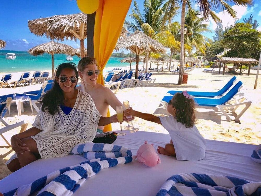 A family of three clinks their glasses together as they sit together in a covered cabana, while on a stunning troppical beach.