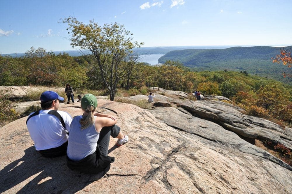 A couple sits upon a giant boulder at the top of Bear Mountain enjoying the perfect fall foliage near NYC.