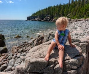 A young boy sits on the rocky coast of Maine with trees in the distance.