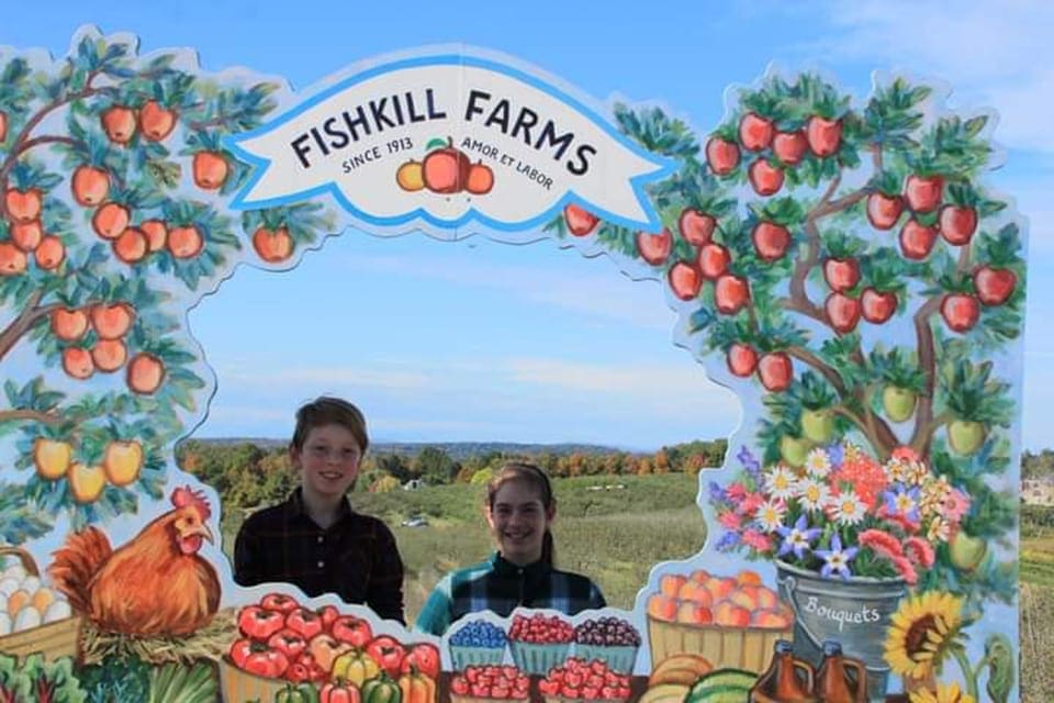 Two kids stand behind a cardboard cutout photograph display featuring apples, pumpkins, and sunflowers at Fishkill Farms, one of the best places for apple picking with kids around NYC.