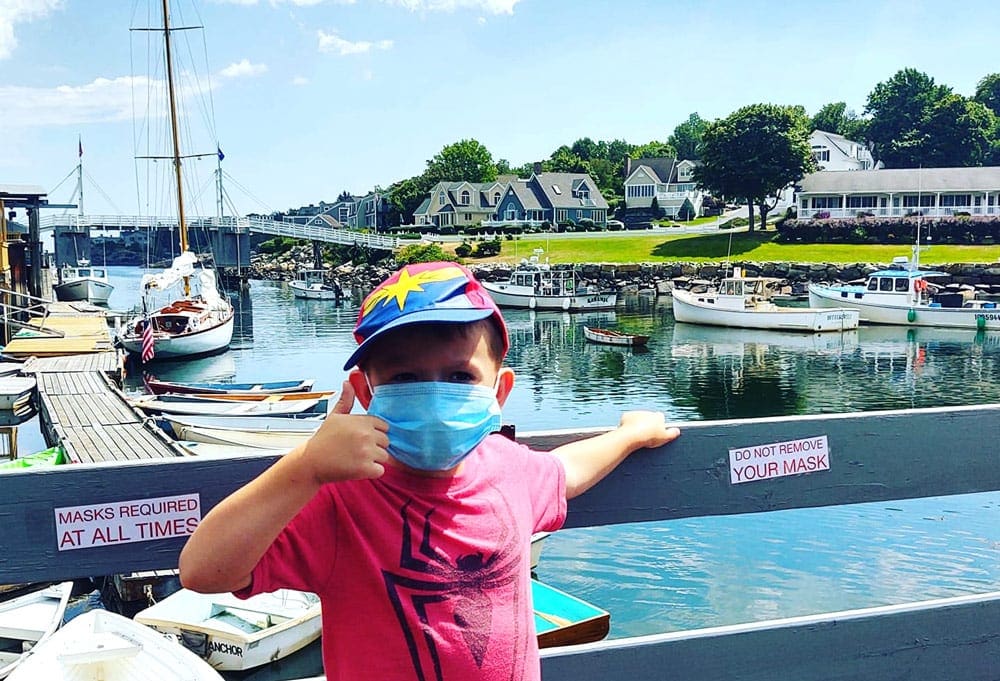 A young boy wearing a mask gives a thumbs up on a bridge in Ogunquit, behind him lies a harbor filled with small boats, as well as historic buildings and large green spaces.