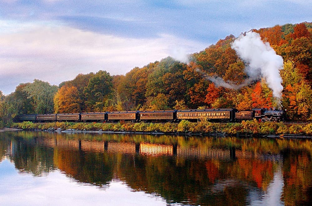 An Essex Steam Train goes full steam ahead along a glimmering lake on a bright red fall day, a great option for a fall foliage train ride in the Northeast.