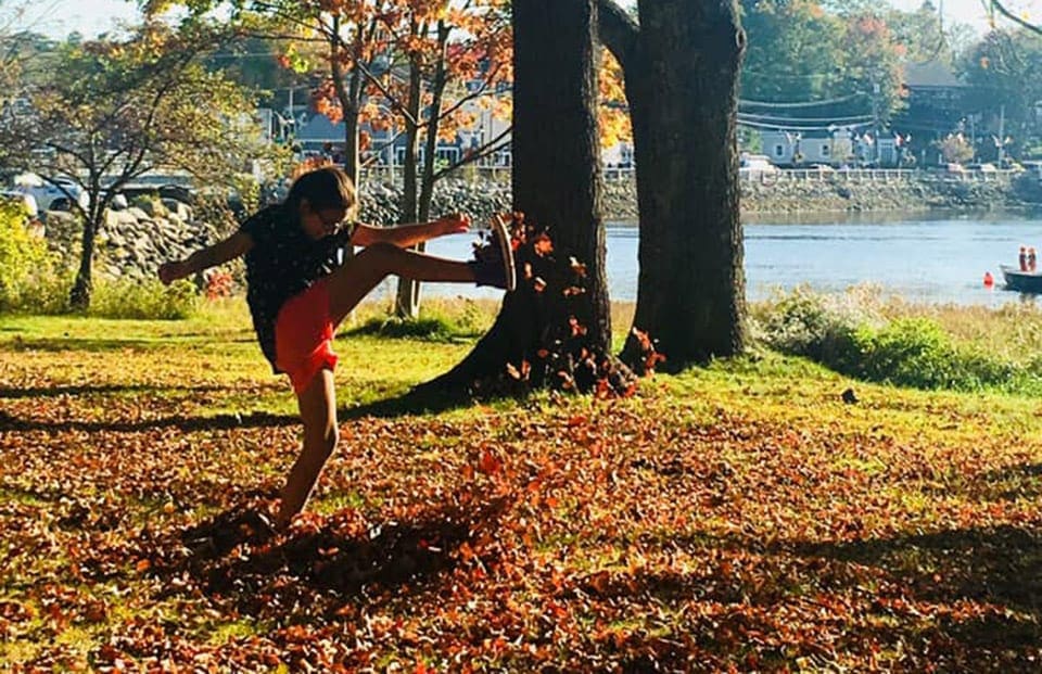 A young child kicks up a lot of fall leaves from a large pile on an autumn day in Maine, one of the best places to see fall colors in the US for families.