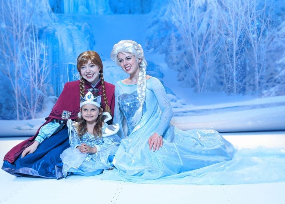 A young girl wearing a princess dress poses with Anna and Elsa of Frozen abroad a Disney Cruise.