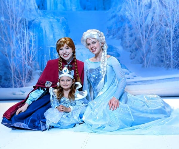 A young girl wearing a princess dress poses with Anna and Elsa of Frozen abroad a Disney Cruise.