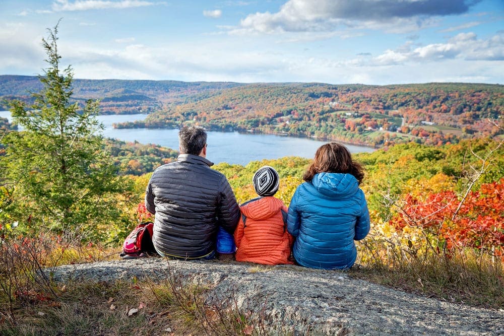 A family of three sits upon a large boulder overlooking a coloful autumn display in New England.