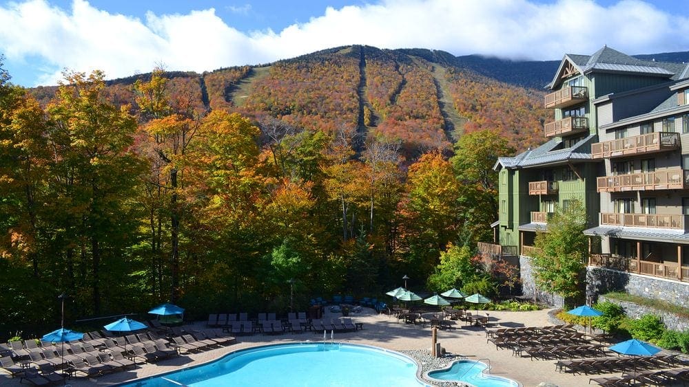 On an autumn day, an outdoor pool shines next to buildings on the grounds of the Lodge at Spruce Peak, with fall hues and ski trails in the background.