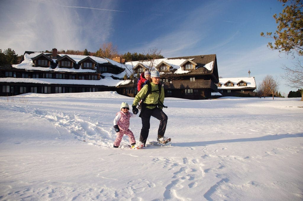 An adult showshoes while carrying one small child on their back and holding the hand of another while they explore the snowy grounds of the Trapp Family Lodge.