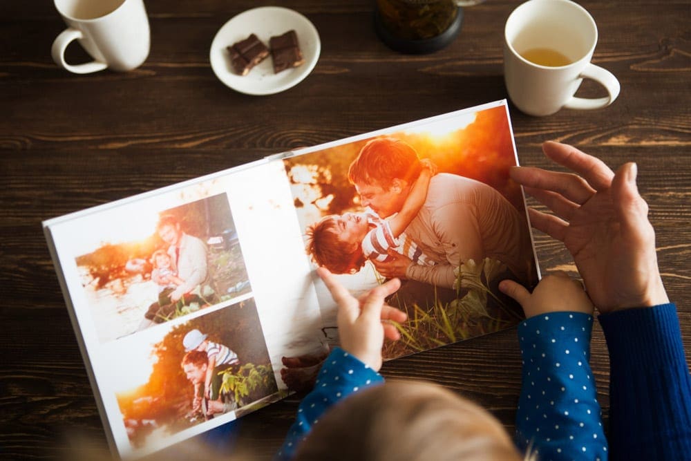 An adult and young baby look at picture in a family photo book, coffee mug and chocolates also featured.