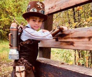 A young boy wearing a Steampunk costume leans against a fence.