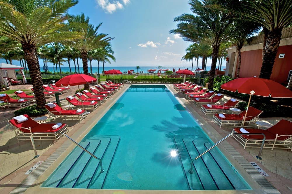 A large pool surrounded by iconic red chairs and umbrellas at The Acqualina Resort.