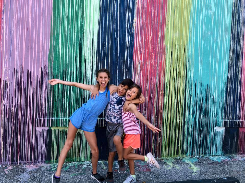 Three kids huddle together, all smiling, in front of a colorful dripping paint street art mural in Houston.