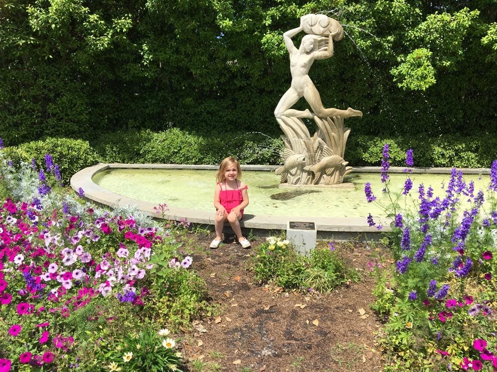 A young girl wearing pink sits next to a fountain surrounded by blooming flowers in City Park.