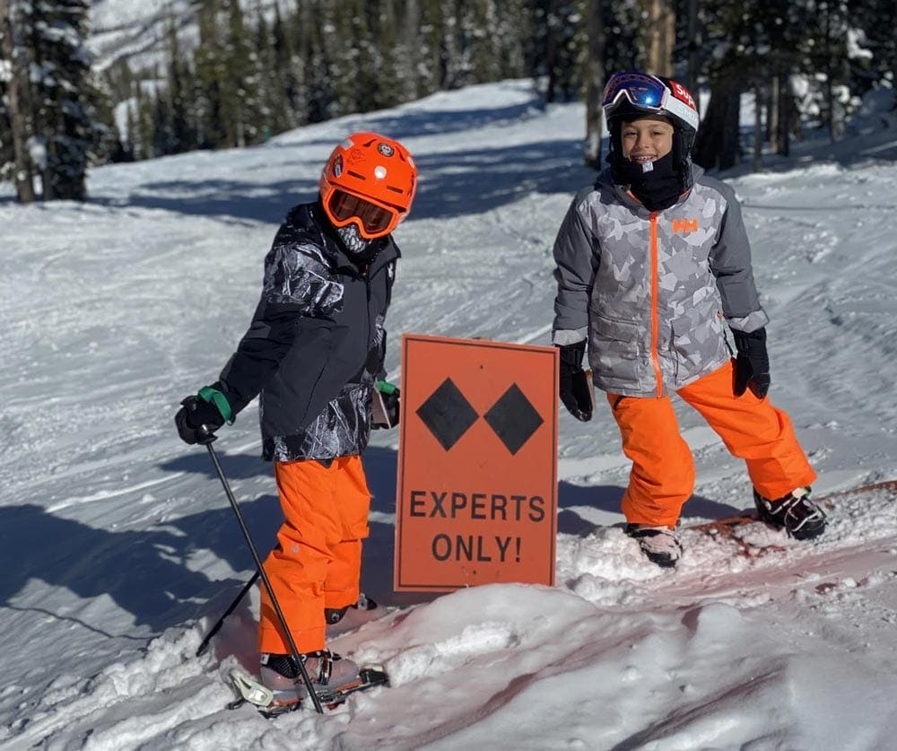 Two boys in full snow gear stand next to a sign reading "Experts Only" while skiing at Beaver Creek.