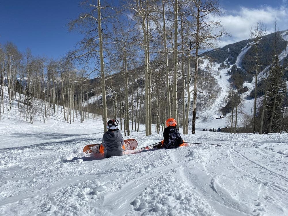 Two young kids sit in the snow, wearing snow gear and snowboards, with an expansive view of mountains at Beaver Creek behind them.