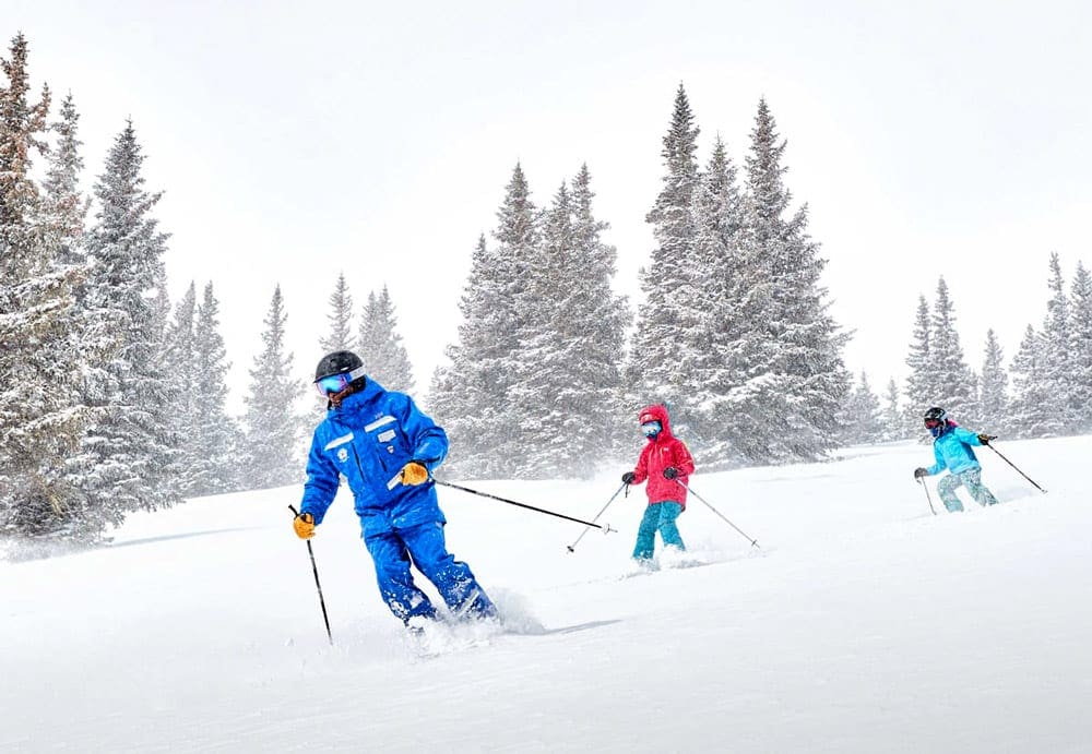 A father wearing a blue suit is followed, on skis, but two kids, one in bright red and one in light blue at Breckenridge Ski Resort.