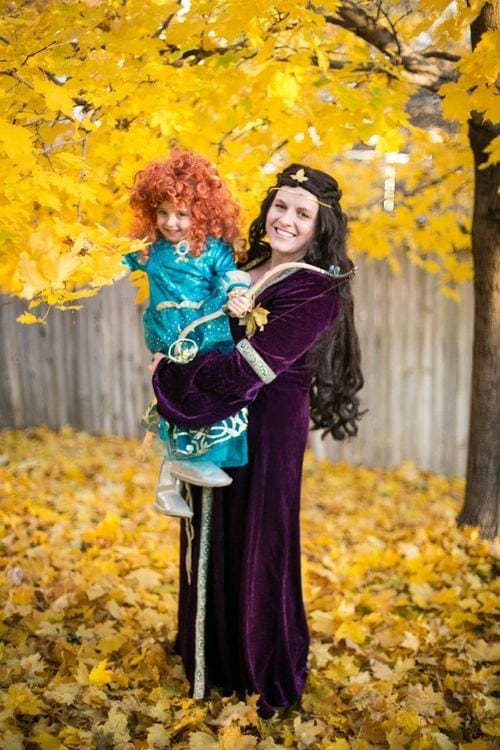 A woman wearing dark purple robes holds a young girl dressed as Merida from the movie Brave.