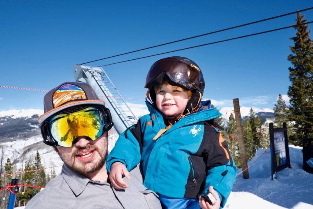 A dad wearing large ski goggles holds his toddler son, ready to ski at Vail.