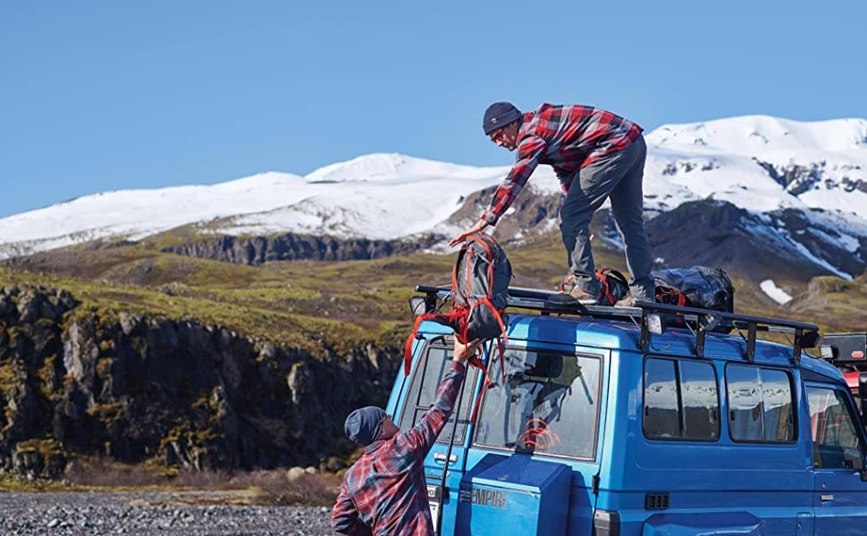 two men with matching red flannel shirts helping one another tie backpacks to the top of a truck. set in a mountain landscape. 